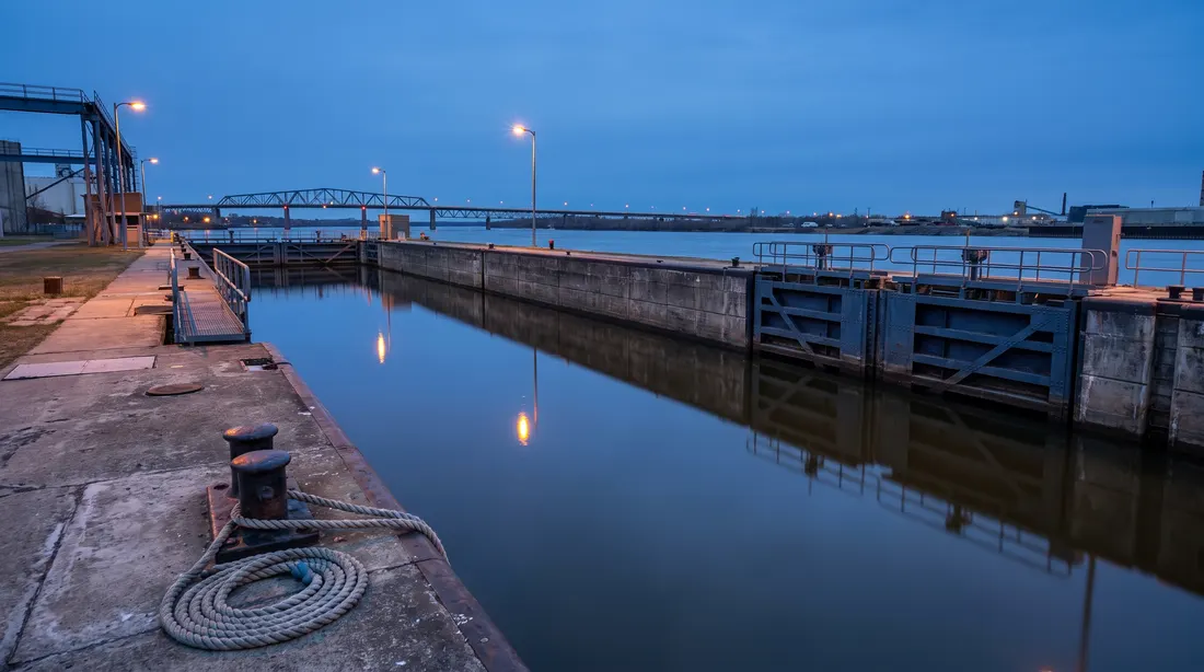Scène générée : une écluse de canal à l'heure bleue, deux portes d'acier fermées au bout d'un long sas rempli d'eau, un pont en treillis d'acier au loin; ce n'est pas une photo du casino.